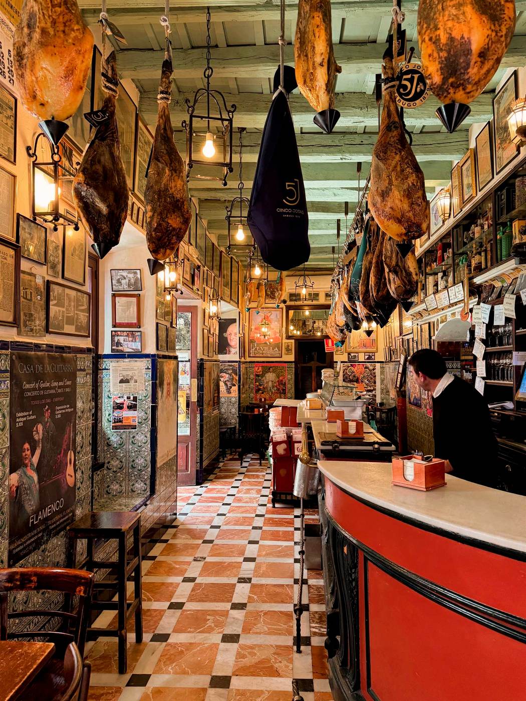 Barra de taberna madrileña al mediodía, vasos de vermú con hielo y naranja, jamón cortado a cuchillo. Luz natural.